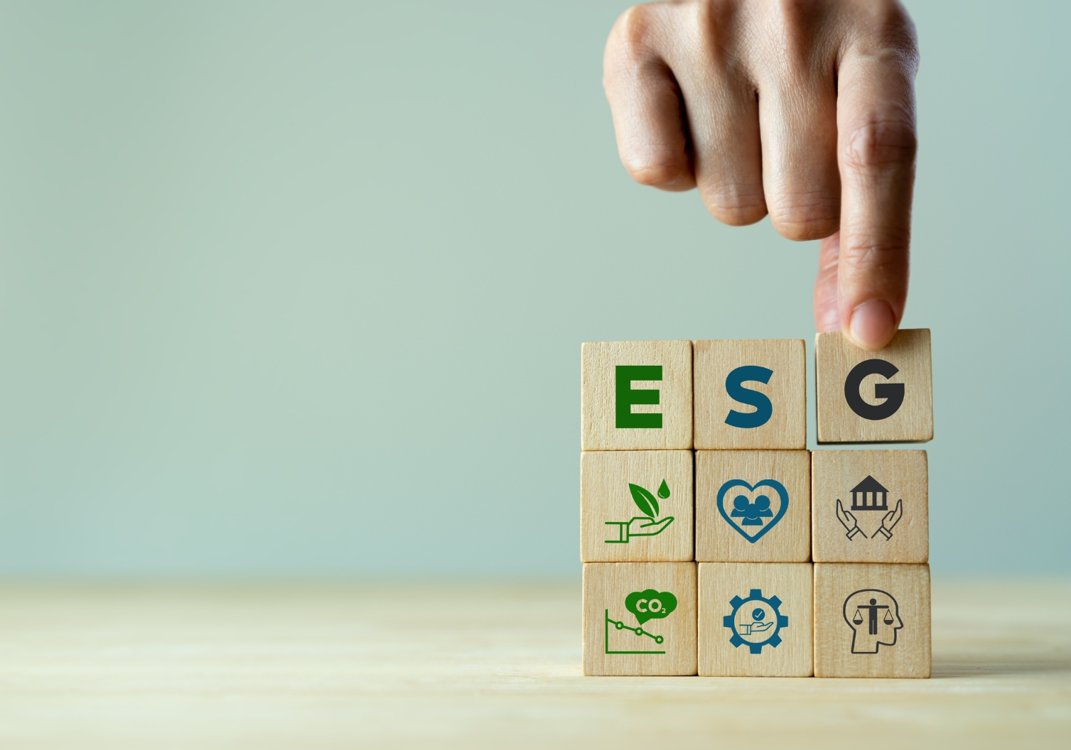 A hand arranges wooden blocks with the letters ESG on them. Below these letters, additional blocks display icons representing environmental, social, and governance themes, such as a leaf, a heart, a factory, and gears.