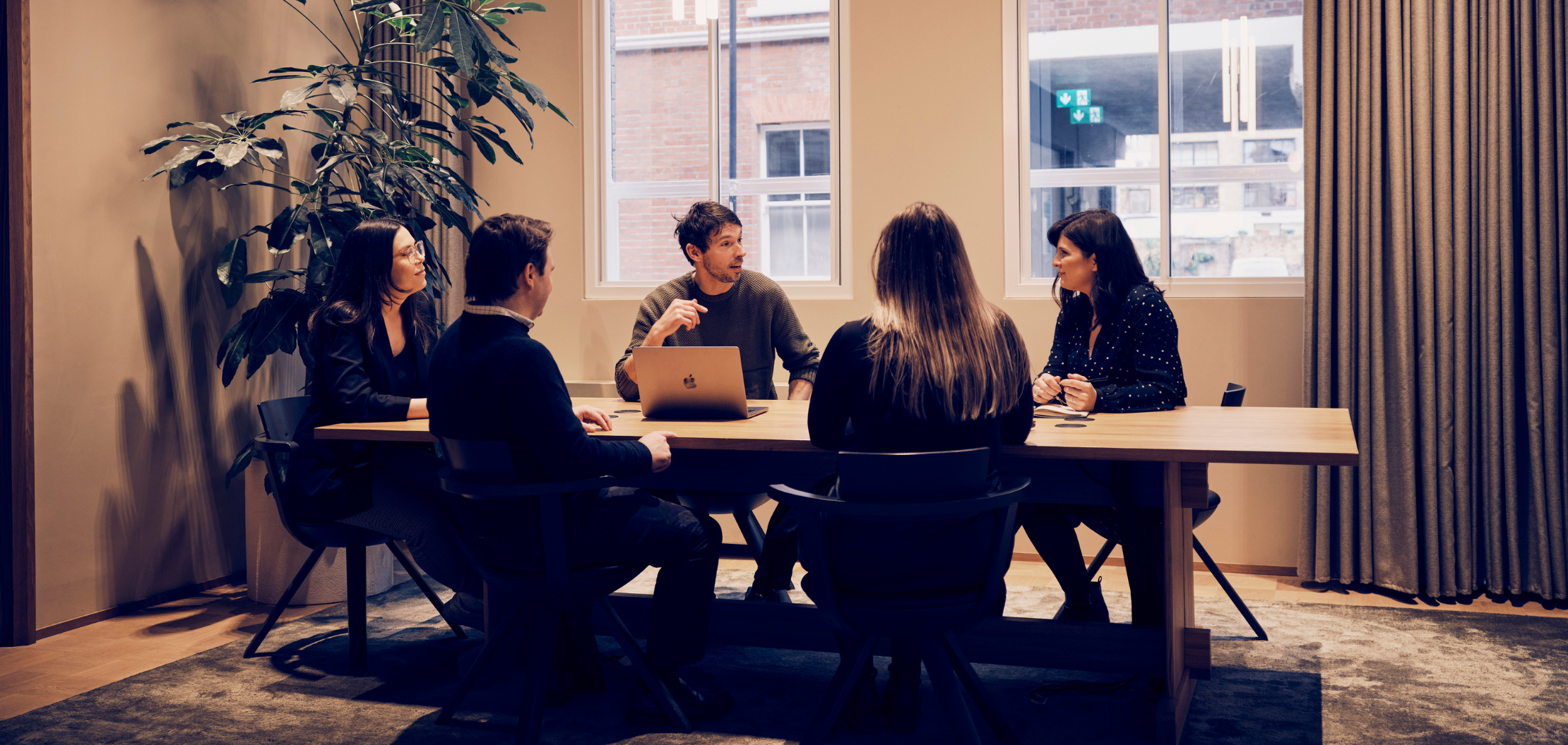 A group of five people sitting around a table in a meeting room of The Think Tank marketing agency with a large plant in the background. One person is speaking and gesturing while the others listen attentively. The room has large windows with views of a brick building outside